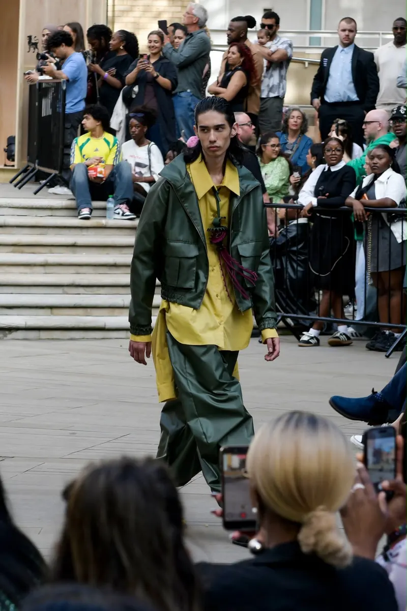 Model in an olive-green jacket over a yellow shirt walking in front of onlookers