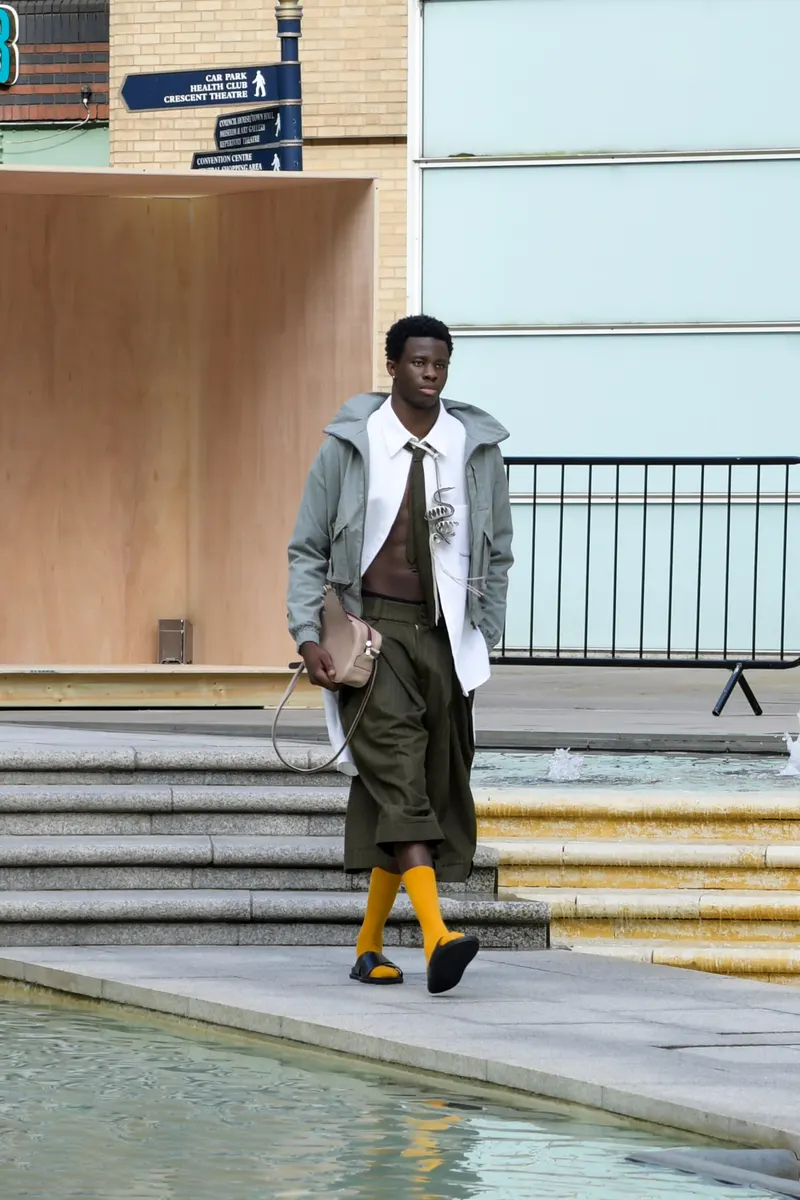 Model in a grey hooded jacket, white shirt and olive shorts walking beside a pool