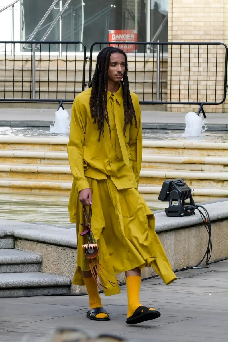 Model in a mustard-yellow shirt and skirt walking past a fountain